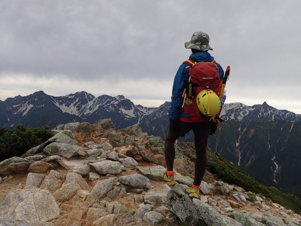 梅雨空の常念岳山頂でモンベルの登山用レインウェアであるトレントフライヤーを着て槍ヶ岳と穂高岳の稜線をバックに記念写真を撮影