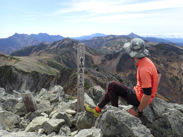 北アルプスの水晶岳の山頂で槍ヶ岳と穂高岳をバックに登山用のゴアッテックスの帽子を被って記念写真を撮影