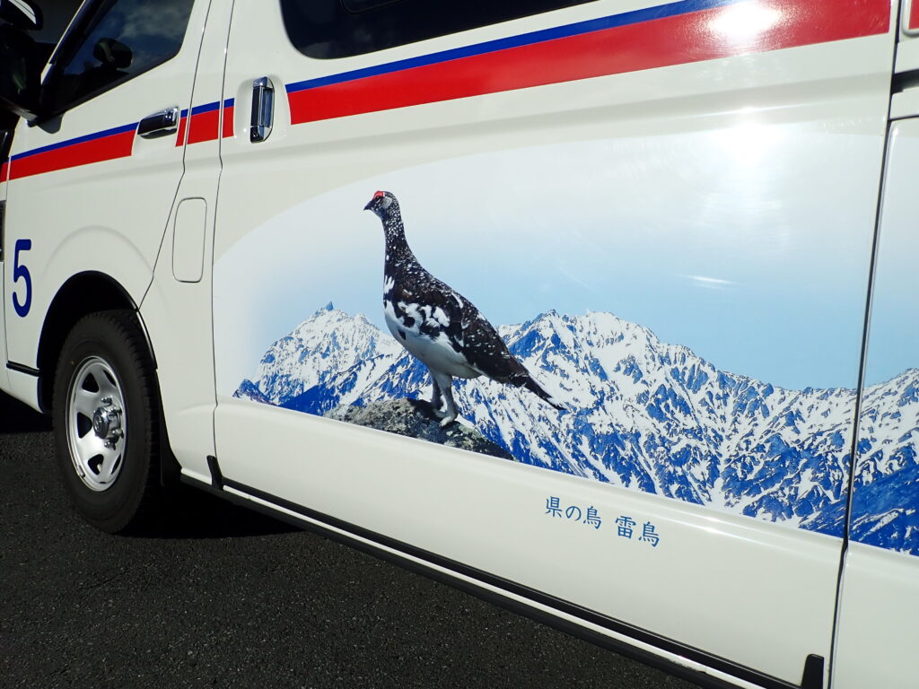 槍ヶ岳から穂高岳の稜線が背景になった雷鳥の写真がプリントされた救急車